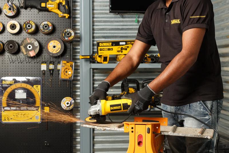 A person wearing gloves uses a Dewalt DW803 angle grinder to cut a metal piece, with sparks flying. Various Dewalt power tools and grinding discs are displayed on a wall and workbench in the background.