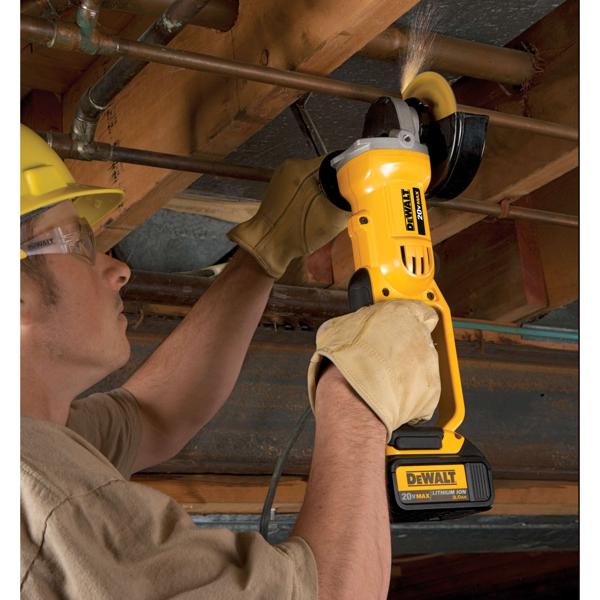 A person wearing protective gloves uses a yellow DEWALT cordless angle grinder to cut a metal pipe under a wooden ceiling. The grinder is battery-powered and produces sparks as it cuts.