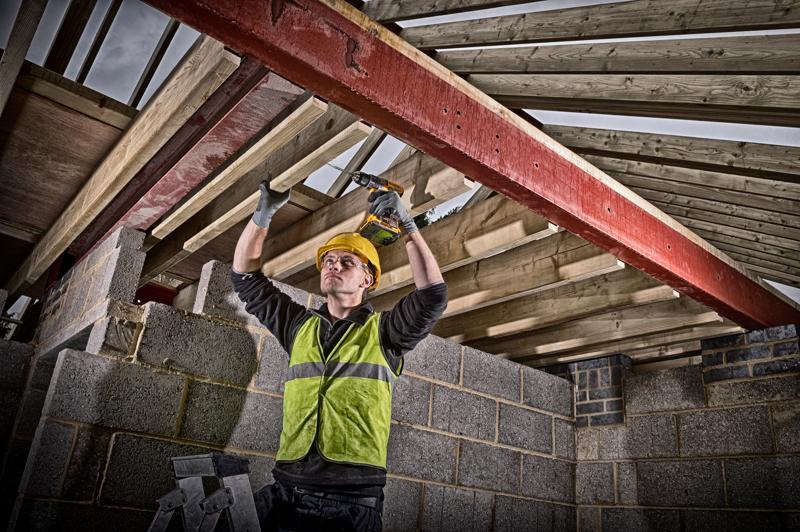 A construction worker wearing a yellow hard hat and high-visibility vest uses a DEWALT cordless drill to secure wooden beams to a red steel support inside a building under construction. The face of the worker is blurred.