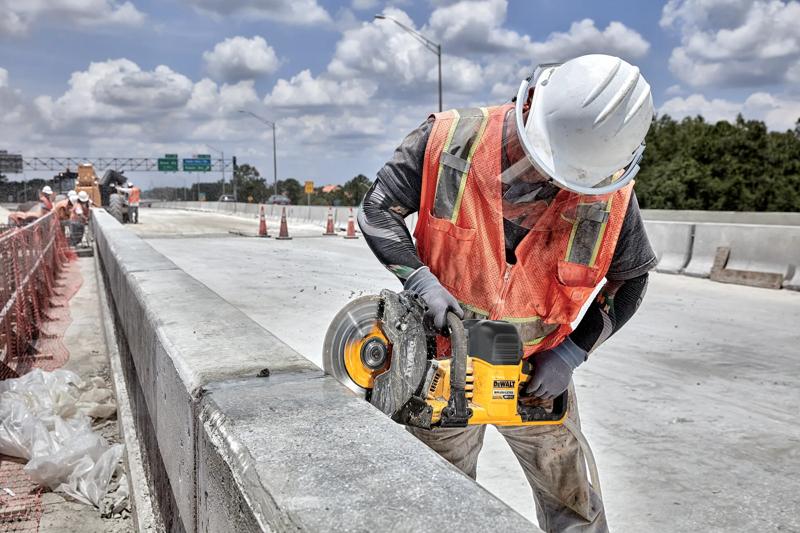 A construction worker wearing a white hard hat and orange safety vest is using a DEWALT cordless cut-off saw (model DCS692B) to cut through a concrete barrier on a road construction site.