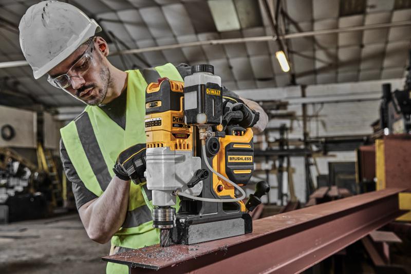 A person wearing a safety helmet and high-visibility vest is operating a DEWALT DCD1624 brushless magnetic drill on a steel beam inside an industrial workshop.