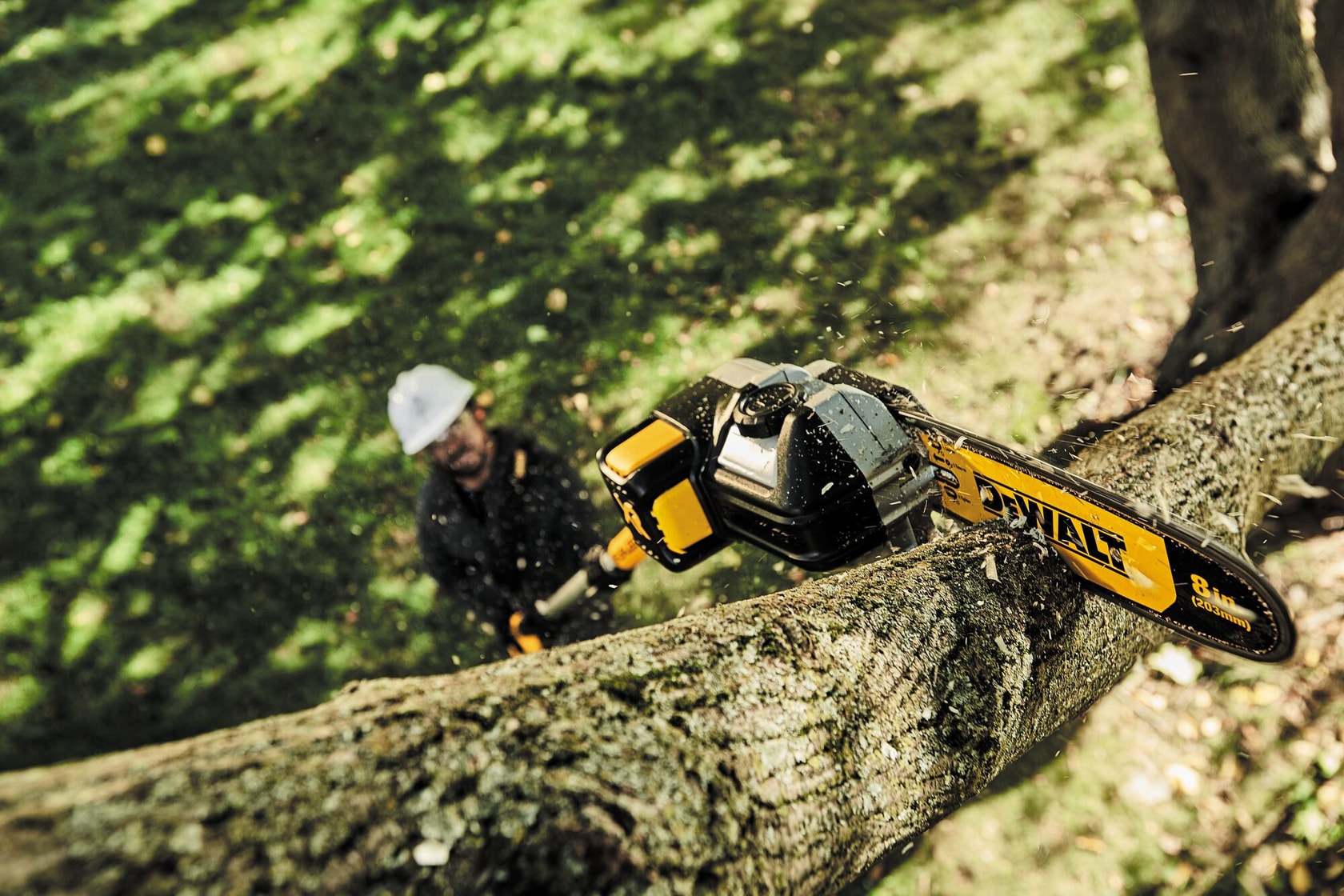 A DEWALT pole saw model DCPS620B cutting a thick tree branch outdoors. The saw is in action, creating wood chips and dust. A person, whose face is blurred, is operating the tool beneath the branch.