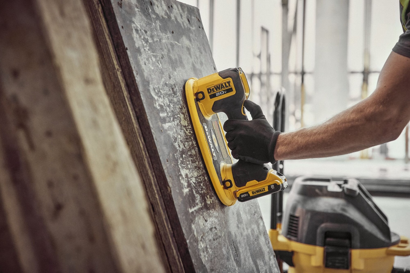 A person's gloved hand using a DEWALT 20V cordless handheld sander on a large, weathered wooden surface, with another DEWALT tool in the background.